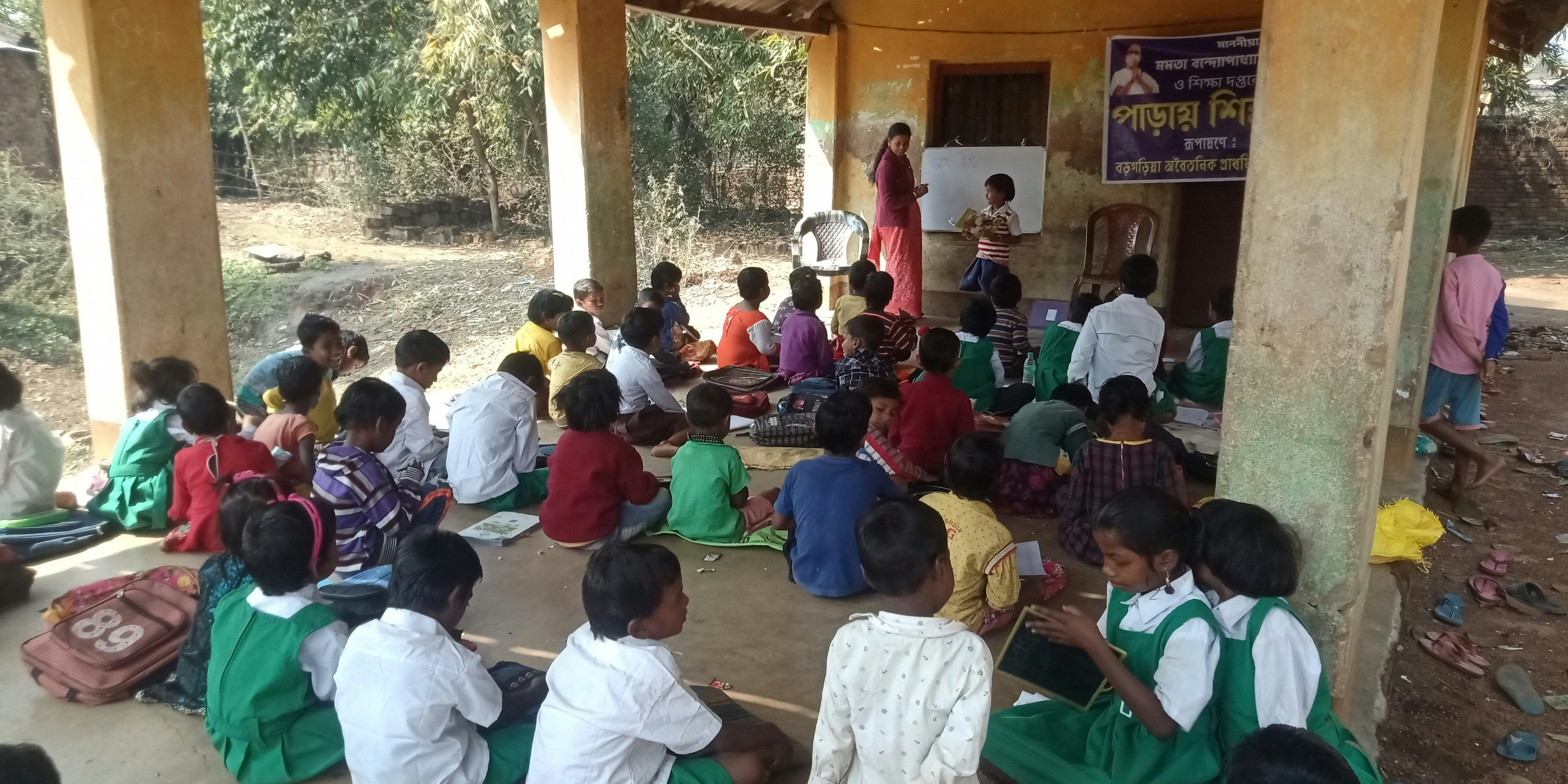 Children sitting in rows at wooden desks in an open-air classroom, teacher writing on blackboard