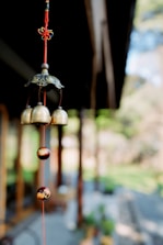 A close-up of a wind chime hanging from a red string. The wind chime consists of several brass bells and ornamental beads hanging vertically. In the blurred background, there appears to be an outdoor setting with vegetation and wooden structures.