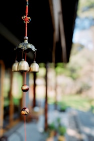 A close-up of a wind chime hanging from a red string. The wind chime consists of several brass bells and ornamental beads hanging vertically. In the blurred background, there appears to be an outdoor setting with vegetation and wooden structures.