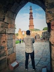 A person stands with their back to the camera, looking at a tall, ancient tower framed by the stone archway of a historic site. The architecture showcases intricate designs, with weathered stone pathways leading to the structure. Other visitors wander the site, capturing the grandeur of the setting.