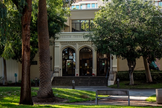 A building facade with the words 'Fuller Theological Seminary' above a set of arched doors, surrounded by lush green trees and neatly manicured lawns. The entrance is accessed by a short staircase, with a bench and pathway leading to it.