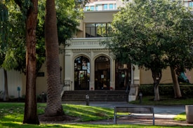 A building facade with the words 'Fuller Theological Seminary' above a set of arched doors, surrounded by lush green trees and neatly manicured lawns. The entrance is accessed by a short staircase, with a bench and pathway leading to it.