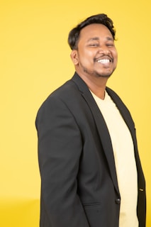 A person is smiling broadly while wearing a black blazer over a yellow shirt. The background is a solid bright yellow, adding a vibrant feel to the image.
