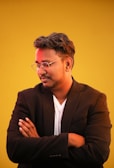A person is standing against a bright yellow background, wearing a black blazer over a white shirt. They are facing slightly to the side with arms crossed, looking down thoughtfully. They have short, styled hair and are wearing glasses.