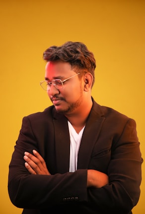 A person is standing against a bright yellow background, wearing a black blazer over a white shirt. They are facing slightly to the side with arms crossed, looking down thoughtfully. They have short, styled hair and are wearing glasses.