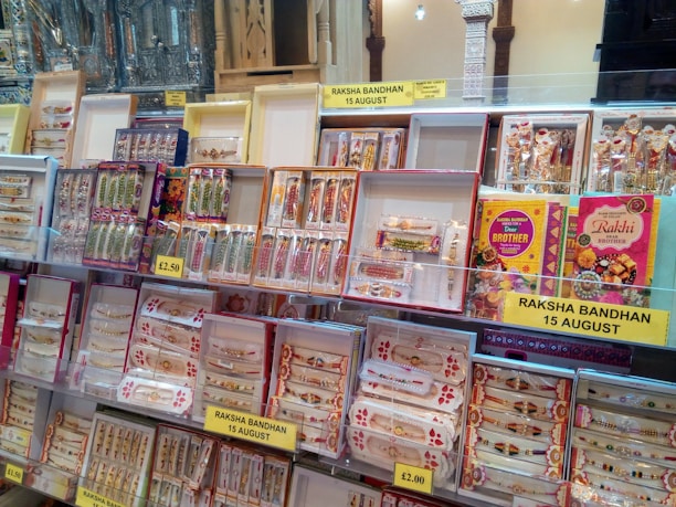 A display of colorful rakhi bracelets arranged in various boxes and packages, positioned on shelves in a store. Labels indicate pricing and the occasion of Raksha Bandhan on August 15th. The display includes decorative elements and traditional patterns, possibly aimed at celebrating the Indian festival.