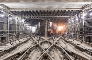 An underground train track with multiple rail lines converging and diverging. Workers wearing orange safety vests are visible in the distance, engaged in construction or maintenance activities. The area is illuminated by artificial lights hanging from the ceiling, and numerous cables and pipes run along the walls and ceiling. The scene is industrial, with a focus on the infrastructure and complexity of the railway system.