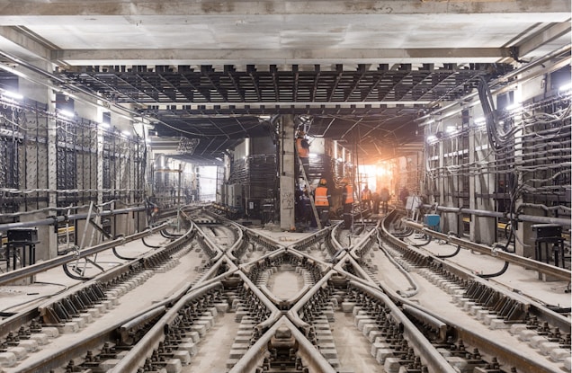 An underground train track with multiple rail lines converging and diverging. Workers wearing orange safety vests are visible in the distance, engaged in construction or maintenance activities. The area is illuminated by artificial lights hanging from the ceiling, and numerous cables and pipes run along the walls and ceiling. The scene is industrial, with a focus on the infrastructure and complexity of the railway system.