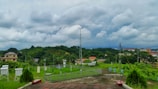 A lush, green landscape with various types of vegetation, including trees and shrubs, is spread out across the scene. Buildings with colorful rooftops are interspersed within the greenery. A tall communication tower and several small meteorological instruments are present in the foreground along a paved path. The sky is filled with thick, dramatic clouds, hinting at an impending storm.