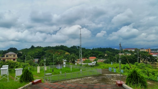 A lush, green landscape with various types of vegetation, including trees and shrubs, is spread out across the scene. Buildings with colorful rooftops are interspersed within the greenery. A tall communication tower and several small meteorological instruments are present in the foreground along a paved path. The sky is filled with thick, dramatic clouds, hinting at an impending storm.