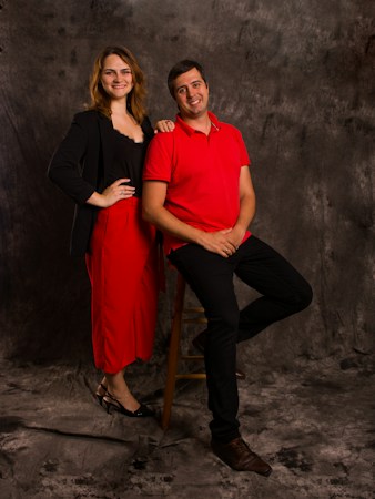A man and a woman are posing together against a textured gray backdrop. The man is seated on a stool, wearing a red shirt and black pants, smiling. The woman is standing beside him, wearing a black blazer over a red outfit and black heels, with one hand on her hip and the other resting on the man's shoulder. Both are smiling and appear casual and relaxed.