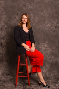 A person with light brown hair is seated on a red stool against a textured grey backdrop. They are wearing a black blazer, a black top, and vibrant red pants, paired with black heels. The pose is relaxed, and the expression is calm and composed.