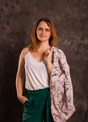 A woman stands confidently against a textured dark gray background, wearing a white sleeveless top with lace trim and green high-waisted trousers. She holds a floral patterned pink fabric over her shoulder, while her hand rests casually in her pocket. Her expression is relaxed and self-assured.