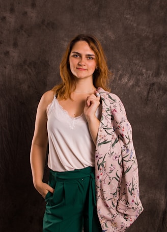 A woman stands confidently against a textured dark gray background, wearing a white sleeveless top with lace trim and green high-waisted trousers. She holds a floral patterned pink fabric over her shoulder, while her hand rests casually in her pocket. Her expression is relaxed and self-assured.