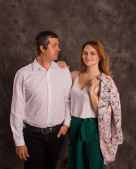 A man and a woman stand closely side by side against a dark textured backdrop. The man, wearing a white shirt and black pants, appears to be looking at the woman. The woman, with light brown hair, is smiling and wearing a white top and green pants, with a floral jacket draped over her shoulder.