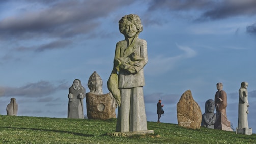 A stone sculpture of a person holding a bird stands prominently on a grassy hill. Several other sculptures and figures are scattered in the background, creating a surreal landscape under a partly cloudy sky. A person wearing a coat and carrying a bag walks among the sculptures, adding a sense of scale to the scene.