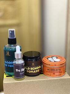 The image depicts a collection of hair care products, including a spray bottle labeled 'Lagoon Hair Tonic', a small bottle labeled 'Pre Style', and jars of 'Old School Pomade' and 'Murray's Superior Hair Dressing Pomade'. These items are arranged on a plain box with a blurred background.