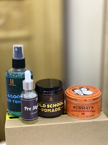 The image depicts a collection of hair care products, including a spray bottle labeled 'Lagoon Hair Tonic', a small bottle labeled 'Pre Style', and jars of 'Old School Pomade' and 'Murray's Superior Hair Dressing Pomade'. These items are arranged on a plain box with a blurred background.