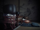 A traditional Japanese tatami room showcasing a large bronze singing bowl placed on an ornate wooden stand with a cushion. Nearby, there is a smaller bowl and a decorative wooden box or altar. The backdrop includes a serene mountain landscape mural, lending an atmosphere of tranquility. Additional items include a meditation cushion or pad and a wooden instrument typically associated with Buddhism.