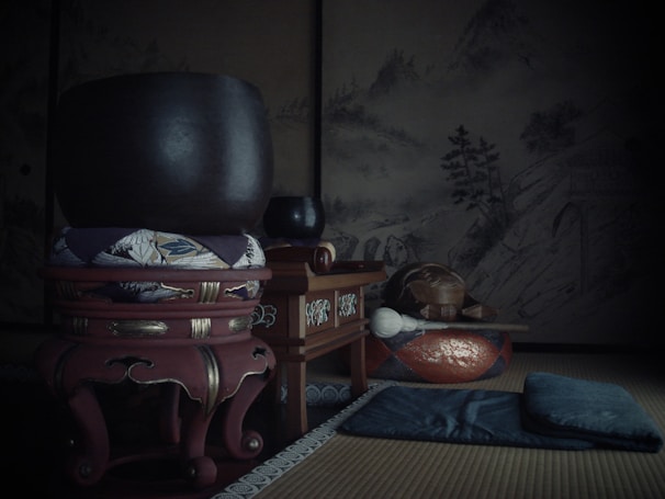 A traditional Japanese tatami room showcasing a large bronze singing bowl placed on an ornate wooden stand with a cushion. Nearby, there is a smaller bowl and a decorative wooden box or altar. The backdrop includes a serene mountain landscape mural, lending an atmosphere of tranquility. Additional items include a meditation cushion or pad and a wooden instrument typically associated with Buddhism.