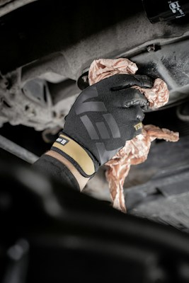 A gloved hand is wiping or cleaning a mechanical part, possibly an engine or a machine, using a crumpled cloth or paper. The setting appears industrial or mechanical, with a focus on maintenance.