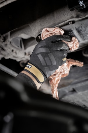 A gloved hand is wiping or cleaning a mechanical part, possibly an engine or a machine, using a crumpled cloth or paper. The setting appears industrial or mechanical, with a focus on maintenance.