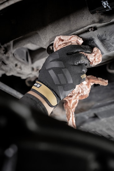 A gloved hand is wiping or cleaning a mechanical part, possibly an engine or a machine, using a crumpled cloth or paper. The setting appears industrial or mechanical, with a focus on maintenance.