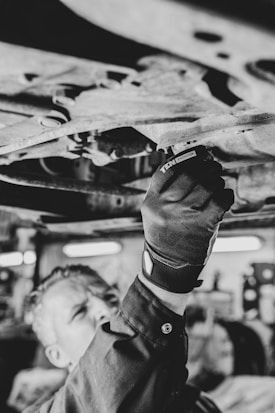 A person is working underneath a vehicle, using a tool to adjust or repair part of the vehicle's undercarriage. The image is in black and white, focusing on the gloved hand and the tool.