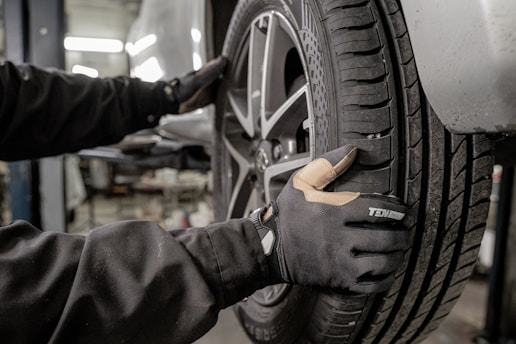 A person wearing protective gloves is handling a car tire. The tire is mounted on a wheel with a metallic rim, and the scene appears to be in a workshop or garage environment, with equipment visible in the blurred background.