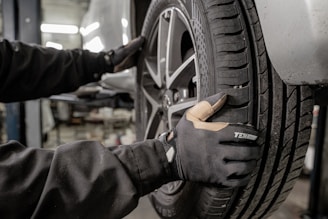 A person wearing protective gloves is handling a car tire. The tire is mounted on a wheel with a metallic rim, and the scene appears to be in a workshop or garage environment, with equipment visible in the blurred background.
