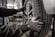 A person wearing protective gloves is handling a car tire. The tire is mounted on a wheel with a metallic rim, and the scene appears to be in a workshop or garage environment, with equipment visible in the blurred background.