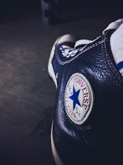A close-up of a black Converse sneaker focusing on the iconic round logo with a blue star and red text. The shoe appears worn, adding a vintage or retro feel.
