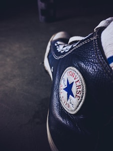 A close-up of a black Converse sneaker focusing on the iconic round logo with a blue star and red text. The shoe appears worn, adding a vintage or retro feel.