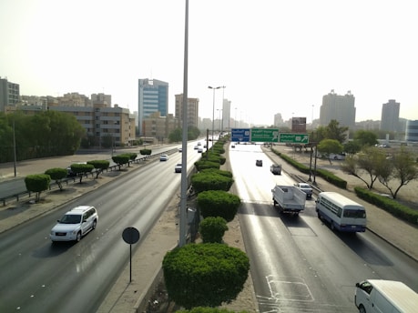 A busy urban highway with multiple vehicles including cars, a bus, and a van. The road is flanked by greenery and neatly trimmed trees. High-rise buildings and skyscrapers are visible in the background under a clear sky, and road signs point towards Kuwait City and other destinations.