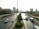 A busy urban highway with multiple vehicles including cars, a bus, and a van. The road is flanked by greenery and neatly trimmed trees. High-rise buildings and skyscrapers are visible in the background under a clear sky, and road signs point towards Kuwait City and other destinations.