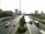 A busy urban highway with multiple vehicles including cars, a bus, and a van. The road is flanked by greenery and neatly trimmed trees. High-rise buildings and skyscrapers are visible in the background under a clear sky, and road signs point towards Kuwait City and other destinations.