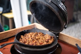 A cast iron skillet filled with shredded meat sits inside an open grill with steam rising, indicating the food is hot and freshly cooked. The setup is on a wooden table, and the surroundings suggest an outdoor setting.