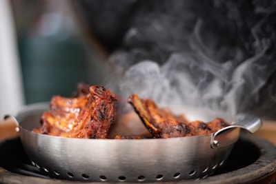 A steaming rack of grilled ribs sits in a metal colander, emitting smoke. The ribs have a charred exterior with a glossy glaze, indicating they are freshly cooked. The background is blurred, with subtle hints of green and black.