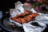 Grilled ribs covered in aluminum foil rest on a barbecue grill. The ribs have a rich, browned appearance, indicative of slow-cooked preparation. Nearby, there's a metal grilling pan with holes, suggesting the potential for cooking vegetables or small items. The background has a slightly blurred view of greenery and flowers, providing an outdoor picnic or garden setting.