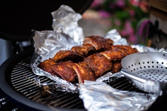 Grilled ribs covered in aluminum foil rest on a barbecue grill. The ribs have a rich, browned appearance, indicative of slow-cooked preparation. Nearby, there's a metal grilling pan with holes, suggesting the potential for cooking vegetables or small items. The background has a slightly blurred view of greenery and flowers, providing an outdoor picnic or garden setting.