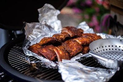 Grilled ribs covered in aluminum foil rest on a barbecue grill. The ribs have a rich, browned appearance, indicative of slow-cooked preparation. Nearby, there's a metal grilling pan with holes, suggesting the potential for cooking vegetables or small items. The background has a slightly blurred view of greenery and flowers, providing an outdoor picnic or garden setting.