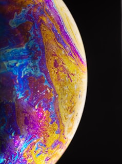A vibrant swirl of colors including blue, purple, and gold, forming intricate patterns across a surface that resembles a planetary body. The image captures the delicate interplay of these colors, evoking a sense of cosmic wonder.