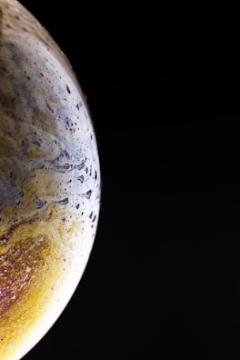 A close-up view of what appears to be a planet with a textured surface, featuring swirls and patches of colors against a pitch-black background. The planet has areas of yellow, brown, and white, mixed with dark speckles and blueish streaks.