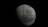 A large, cratered celestial body appears prominently against a pitch-black background. The surface is textured with craters and ridges, indicating an airless, rocky terrain. The lighting creates a stark contrast, highlighting one side and shading the other.