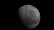 A large, cratered celestial body appears prominently against a pitch-black background. The surface is textured with craters and ridges, indicating an airless, rocky terrain. The lighting creates a stark contrast, highlighting one side and shading the other.