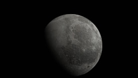 A large, cratered celestial body appears prominently against a pitch-black background. The surface is textured with craters and ridges, indicating an airless, rocky terrain. The lighting creates a stark contrast, highlighting one side and shading the other.