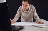 A person leaning over a table, focused on drawing or writing on a large sheet of paper while looking at the screen of an open laptop. Their attire is casual, and they hold a pencil in their right hand. A notebook is placed on the table beside them.