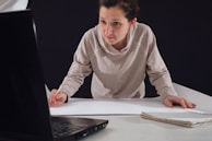 A person leaning over a table, focused on drawing or writing on a large sheet of paper while looking at the screen of an open laptop. Their attire is casual, and they hold a pencil in their right hand. A notebook is placed on the table beside them.