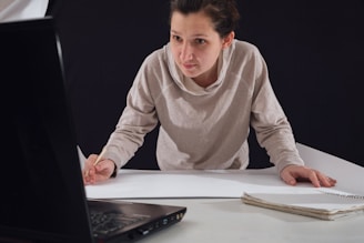 A person leaning over a table, focused on drawing or writing on a large sheet of paper while looking at the screen of an open laptop. Their attire is casual, and they hold a pencil in their right hand. A notebook is placed on the table beside them.