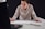 A person leaning over a table, focused on drawing or writing on a large sheet of paper while looking at the screen of an open laptop. Their attire is casual, and they hold a pencil in their right hand. A notebook is placed on the table beside them.